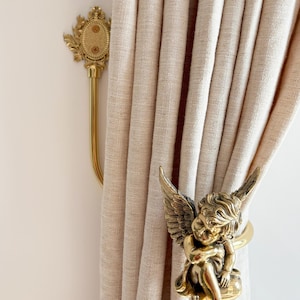 Puede incluir: Recogedor de cortina dorado con un querubín alado sentado, sujetando una cortina beige. El querubín tiene detalles finos. También se ve un soporte de barra de cortina dorado a juego.