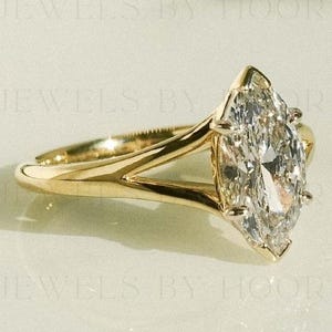 Puede incluir: Un anillo de compromiso con diamante talla marquesa y banda dividida. El anillo presenta un diamante grande y transparente engastado en una banda de oro. La banda tiene un diseño dividido cerca del engaste del diamante.