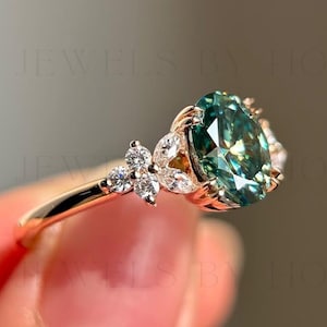 May include: A gold ring featuring an oval, teal-colored gemstone as the centerpiece. The ring is adorned with small, clear accent stones and a floral design on the band. The ring is held up by a person's fingers.