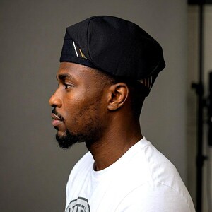 Puede incluir: Una gorra negra con una corona redondeada y estructurada y un borde plano y plegado. El lateral de la gorra presenta un peque&ntilde;o detalle rectangular con finas rayas diagonales en blanco, verde y dorado sobre la tela negra lisa.