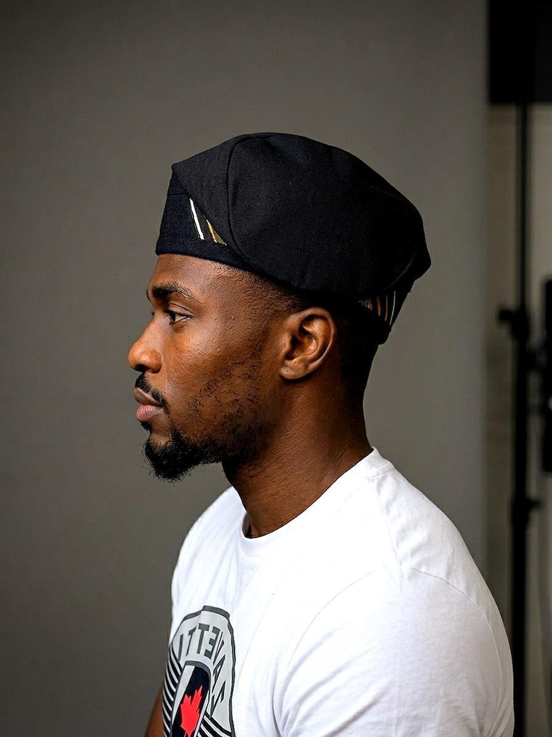 Puede incluir: Una gorra negra con una corona redondeada y estructurada y un borde plano y plegado. El lateral de la gorra presenta un peque&ntilde;o detalle rectangular con finas rayas diagonales en blanco, verde y dorado sobre la tela negra lisa.
