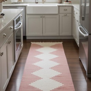 Op de afbeelding: Een rechthoekige loper met een geometrisch patroon in een keuken. De loper heeft een herhalend ontwerp van rode en witte zigzags. De keuken heeft witte kasten, een roestvrijstalen koelkast en een groot raam.