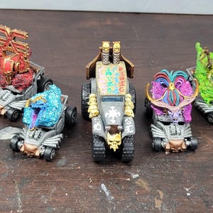 May include: Five custom-painted toy cars with unique, fantasy-themed designs. Each car features intricate details, vibrant colors, and distinct character-like toppers. The cars are set against a neutral background.