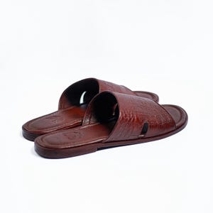 Puede incluir: Un par de sandalias deslizantes de cuero marrón con un patrón texturizado similar al de un cocodrilo. Las sandalias de punta abierta tienen una correa en la parte superior y un pequeño recorte en el lateral. Las sandalias están sobre un fondo blanco.