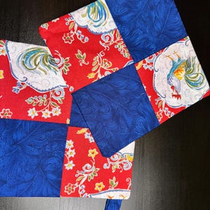 Könnte beinhalten: Zwei rote und blaue gequiltete Topflappen mit einem Hahn- und Blumenmuster. Die Topflappen haben eine Schlaufe zum Aufhängen.