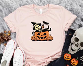 May include: Pale pink t-shirt with a Halloween graphic. The design depicts a skeleton in a witch's hat, lounging on a carved pumpkin, with a bat and smaller pumpkins. White trainers and a skull are also visible in the image.