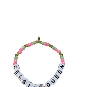 May include: A pink and green beaded bracelet with the words "Celsius Queen" spelled out in white letter beads.