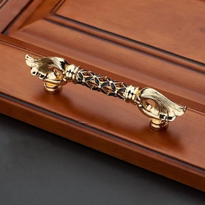 Pode incluir: Puxador de armário dourado ornamentado com um design floral detalhado. A pega apresenta uma secção central com detalhes em preto e termina com um motivo de mão estilizada. Montado numa porta de armário de madeira.