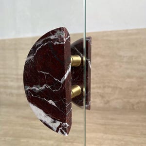 May include: A semi-circular door handle made of deep red marble with prominent white veining. The handle is mounted onto a clear glass panel using two cylindrical, brushed gold-toned metal connectors that secure the stone piece in place.
