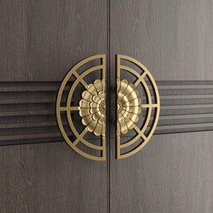 May include: Close-up of a pair of cabinet door handles. Each handle is a half-circle with a floral design in the center, and a geometric pattern around the flower. The handles are gold-colored and mounted on a dark gray wooden cabinet.