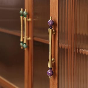 May include: Close-up of a wooden cabinet with glass doors and decorative brass handles. The handles feature a gold finish with purple and green accents, adding a touch of elegance to the furniture.