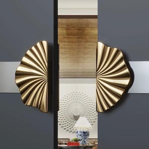 Può includere: Un paio di maniglie dorate a forma di ventaglio. Le maniglie sono montate su una porta grigio scuro con una striscia bianca. Il design presenta una serie di linee curve che si irradiano da un punto centrale, creando un effetto a raggiera. Le maniglie sono un elemento decorativo.
