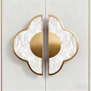 May include: Two decorative cabinet door handles. Each handle features a gold-colored, clover-shaped frame with a mosaic of white, square tiles. A gold-colored, semi-circular piece is in the center of each handle. The handles are mounted on a white cabinet door.