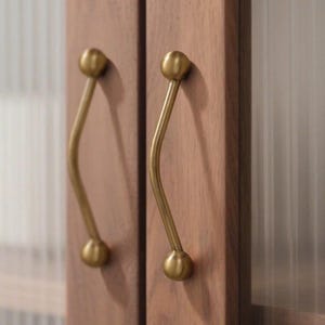 May include: Close-up of two wooden cabinet doors featuring brass-toned, curved handles. Each handle has a spherical end and a gently curving stem, offering a stylish, contemporary look. The wood grain is visible, adding warmth to the design.