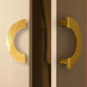 Peut inclure: Deux poignées de placard dorées. L'une a une texture martelée, l'autre est lisse. Les deux sont en forme de croissant et montées sur une porte de placard de couleur claire. Les poignées sont en métal et sont conçues pour une prise en main facile.