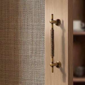 Pode incluir: Uma maçaneta de porta bronzeada com um design texturizado e martelado. A maçaneta é montada em uma porta de madeira clara. O fundo apresenta um painel tecido e texturizado. A maçaneta tem uma barra vertical com um padrão texturizado e barras horizontais em cada extremidade.