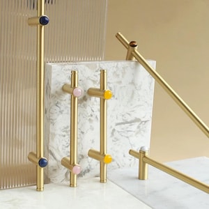 Op de afbeelding: Goudkleurige kastgrepen met kleurrijke, ronde accenten. De grepen hebben een T-bar ontwerp en worden gepresenteerd tegen een marmerachtig oppervlak en een helder, getextureerd paneel. De accenten zijn in tinten blauw, roze, geel en wit.