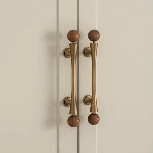 Peut inclure: Deux poignées de porte d'armoire avec une finition en laiton et des détails de boules en bois. Les poignées ont une forme incurvée et allongée et sont montées sur une porte d'armoire de couleur claire. Les boules en bois sont en haut et en bas des poignées.