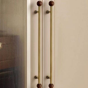 May include: Two cabinet door pulls with a brass finish and dark brown wooden ball ends. The pulls are long and slender, mounted on a cream-coloured cabinet door. The door on the left has vertical ribbed glass.