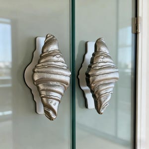 May include: Two silver-colored door handles with a textured, layered design, resembling a croissant shape. Each handle is mounted on a white, scalloped backplate with a black border, attached to a glass door. The handles are a unique decorative element.