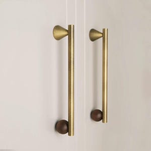 May include: Two cabinet pulls with a brass finish. Each pull has a conical top, a long, straight handle, and a small, round, dark brown wooden knob at the bottom. The pulls are mounted on a white cabinet door.