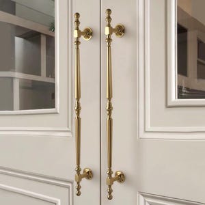May include: Two gold cabinet door handles with an ornate, elongated design. The handles are mounted on a white cabinet door. The handles have a classic, elegant style.