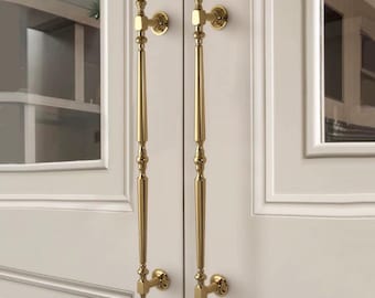 May include: Two gold-colored cabinet door handles with an ornate, elongated design. The handles are mounted on a white cabinet door. The handles have a classic, elegant style.