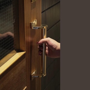 May include: A gold door handle with a curved design is mounted on a wooden door. A hand is reaching for the handle.