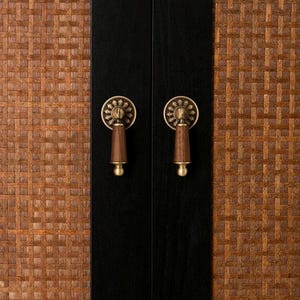 May include: Close-up of a cabinet door with a woven brown texture on the right and black on the left. Two brass-colored handles with wooden pulls are centered on the black doors. The handles have a floral design.