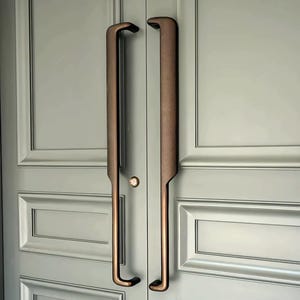 Puede incluir: Primer plano de un par de manijas de puerta de color bronce. Las manijas son largas, verticales y curvas en la parte superior e inferior. Están montadas en una puerta gris claro con paneles empotrados. Un pequeño pomo redondo está centrado.