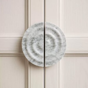 May include: A pair of semi-circular cabinet pulls made of white marble with grey veining. Each pull features a concentric, stepped circular design that creates a ripple effect. The two pieces are mounted together to form a complete circle.