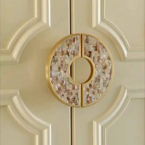 May include: A decorative cabinet pull with a gold-colored frame and a circular design. The center of the pull is filled with a mosaic of iridescent, neutral-toned pieces. The pull is mounted on a cream-colored cabinet door.