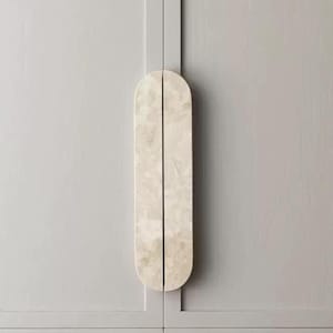 Peut inclure: Une poignée de placard verticale en forme de pilule avec une texture beige clair rappelant la pierre marbrée. La poignée est divisée en son centre par une fine ligne verticale et est fixée sur une porte de placard gris clair au grain de bois visible.