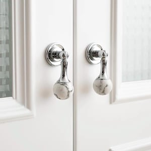 Könnte beinhalten: Zwei weiße Marmor- und Chrom-Schrankknöpfe mit rundem, strukturiertem Design. Die Knöpfe sind an weißen Türen montiert.