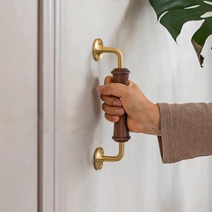 May include: A brass and wood door handle with a curved arm and a wooden grip. The handle is mounted on a white wall.