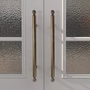 Peut inclure: Gros plan sur une porte blanche avec deux poignées de tirage en laiton antique. Les poignées sont longues, cylindriques et ont des extrémités décoratives en forme de boule. La porte comporte des panneaux de verre à l'aspect texturé et givré.