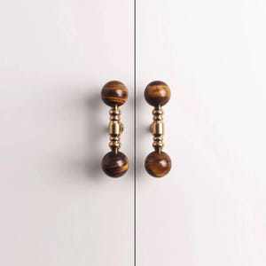 May include: Two cabinet pulls with tiger's eye stone spheres at each end, connected by a gold-coloured metal stem. The pulls are mounted on a white cabinet door. The tiger's eye has brown and gold tones.