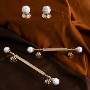 Può includere: Una collezione di ferramenta per mobili con dettagli in metallo dorato e marmo bianco. L'immagine mostra vari pomelli e maniglie, inclusi pomelli rotondi e barre, tutti con un'estetica lussuosa.