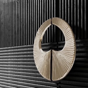Puede incluir: Un tirador de armario ovalado de color dorado con un diseño texturizado en forma de abanico. El tirador está montado sobre una superficie negra ranurada vertical y horizontalmente, creando un contraste de texturas y colores. La ferretería añade elegancia.