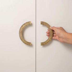 Può includere: Due maniglie per armadietto dorate, a forma di semicerchio, con una superficie strutturata, montate su un'anta bianca. Una maniglia è tenuta in mano. Le maniglie sono un elemento decorativo.