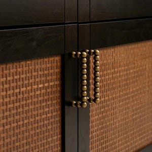 以下が含まれることがあります： 織りラタンパネルと濃い木製フレームを備えたキャビネットドアのクローズアップ。ドアには真鍮製のビーズハンドルが付いています。キャビネットのデザインは、自然な質感とクラシックな美学の融合を示しています。
