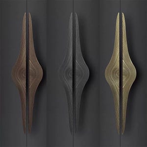 May include: Three decorative cabinet handles in bronze, dark gray, and gold. Each handle has a unique, elongated shape with a central, circular design and a textured, linear pattern. The handles are mounted on a dark gray surface.