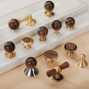 Puede incluir: Una colección de pomos y tiradores para armarios. Los pomos presentan una combinación de madera marrón oscuro y metal dorado. Los diseños incluyen estilos redondos, cilíndricos y de tapa plana, ofreciendo una variedad de opciones.