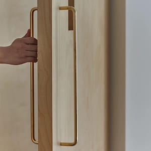 May include: A long, vertical, gold-toned metal door handle with a rectangular profile, mounted on a light-coloured wooden door. The handle features a smooth, continuous design with rounded corners, extending along the edge of the wooden panel.