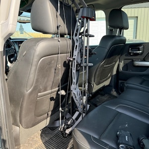 May include: A grey and black compound bow mounted on a headrest in the back seat of a vehicle. A rangefinder is on the seat next to the bow.