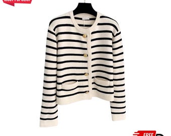 May include: Cream-coloured cardigan with black horizontal stripes. The sweater has a round neckline, gold-coloured buttons, and two front pockets. The sleeves are long, and the cardigan is displayed on a black hanger.