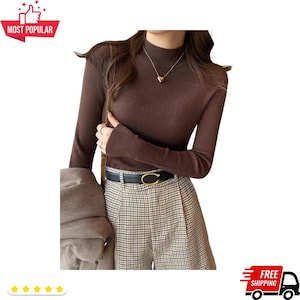 May include: A brown ribbed knit turtleneck sweater. The long-sleeved top is paired with plaid shorts and a black belt with a gold-colored buckle. A heart-shaped pendant hangs from a delicate necklace.