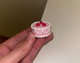 May include: A miniature round cake figurine in a soft pink hue, featuring white rope-style piping along the top and bottom edges, white scalloped detailing on the sides, and three small, glossy red spheres arranged on the top surface.