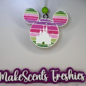 May include: A pink, green, and white Mickey Mouse-shaped ornament with a white silhouette of a castle inside. The ornament has a green bead at the top and the text "Make Scents Freshies" is visible below.