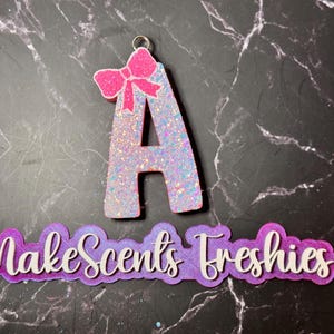 May include: A white glitter letter A with a pink bow on top. The letter is a charm and has a hole at the top for hanging. The background is a dark gray marble surface. The text "MakeScents Freshies" is written in white letters on a purple background.